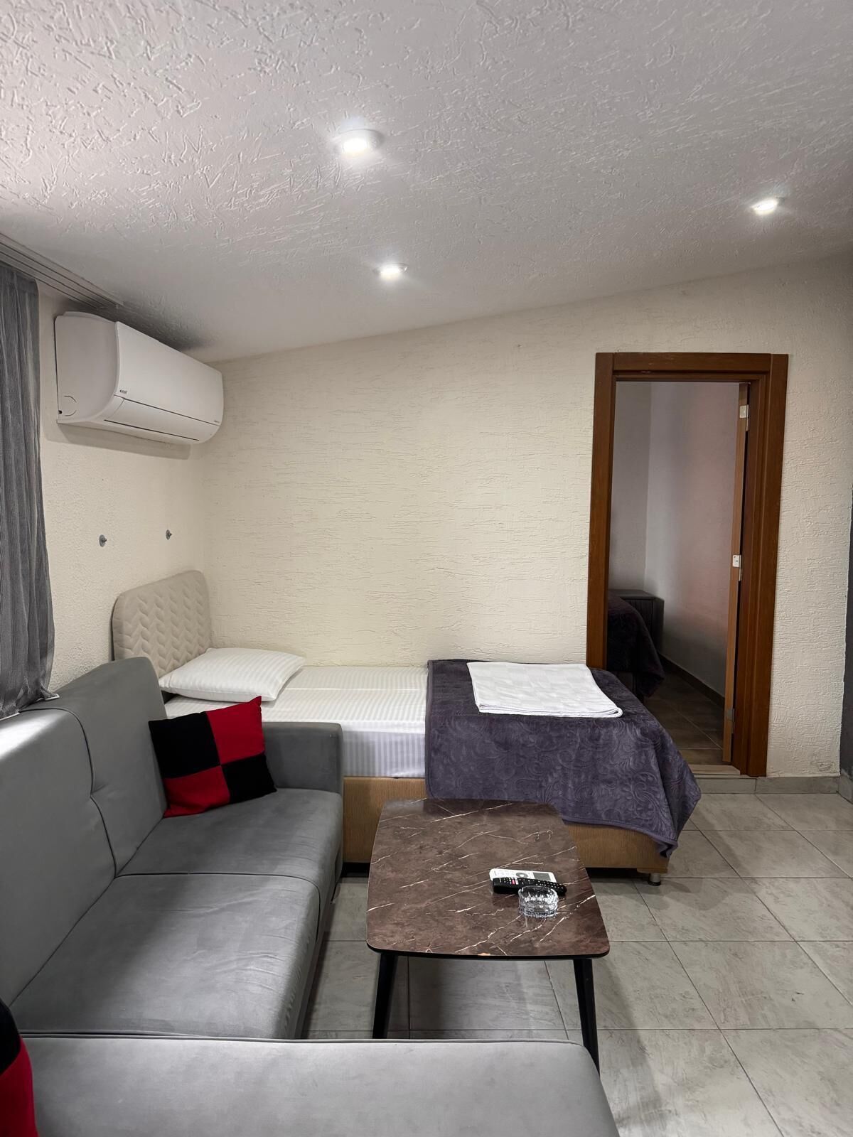Une petite pièce bien éclairée, avec un canapé gris, un lit recouvert d’une couverture violette, une petite table sur laquelle se trouve une télécommande et un appareil de climatisation fixé au mur.