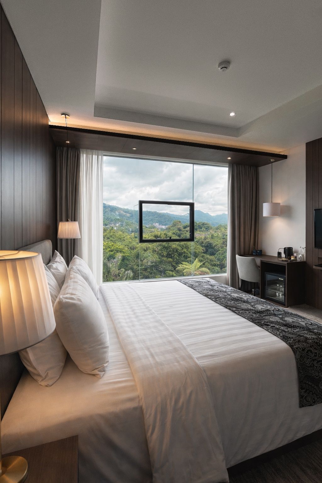 A modern bedroom with a large bed, a desk, and a view of a mountainous landscape.