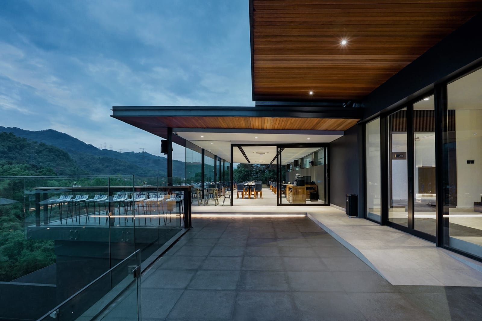 Un edificio moderno con fachada de vidrio, una terraza con asientos al aire libre y vistas a las montañas.