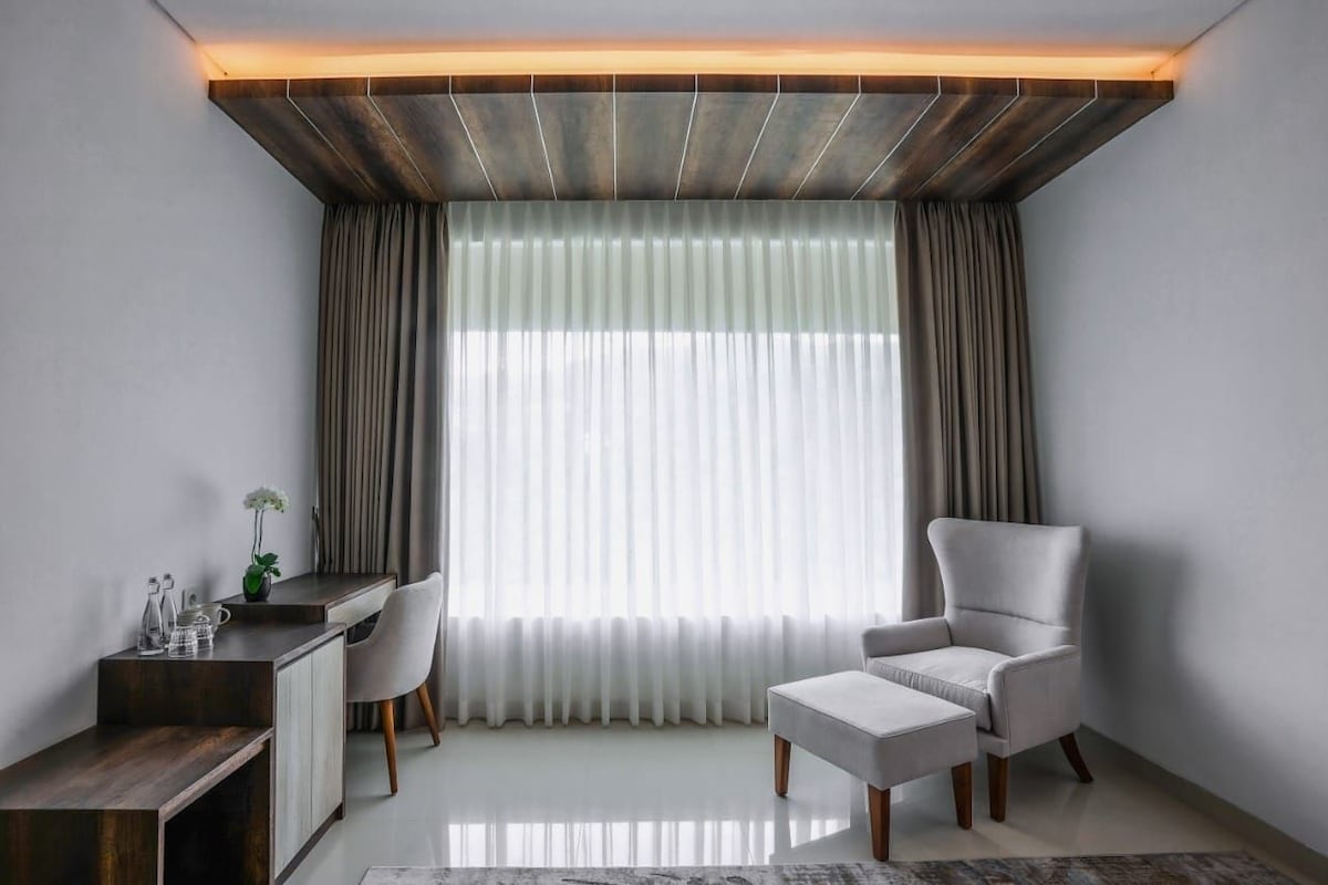 Habitación moderna con escritorio, silla, reposapiés, ventana grande con cortinas y techo de madera.