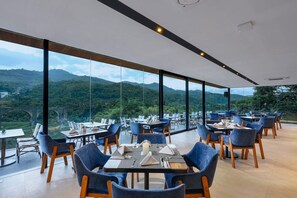 A restaurant with tables and chairs arranged in a spacious area with large windows offering a view of a mountainous landscape. - Forest Hills Hotel Ciwidey (Pasirjambu)