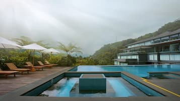 A rooftop infinity pool with wooden loungers and umbrellas, surrounded by lush greenery and a modern building.
