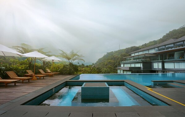 A rooftop infinity pool with wooden loungers and umbrellas, surrounded by lush greenery and a modern building. - Forest Hills Hotel Ciwidey (Pasirjambu)