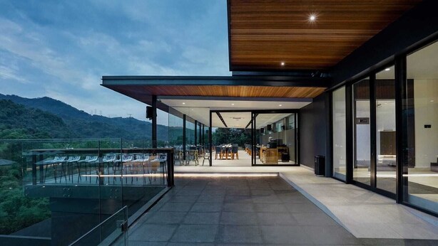 Un edificio moderno con fachada de vidrio, una terraza con asientos al aire libre y vistas a las montañas.