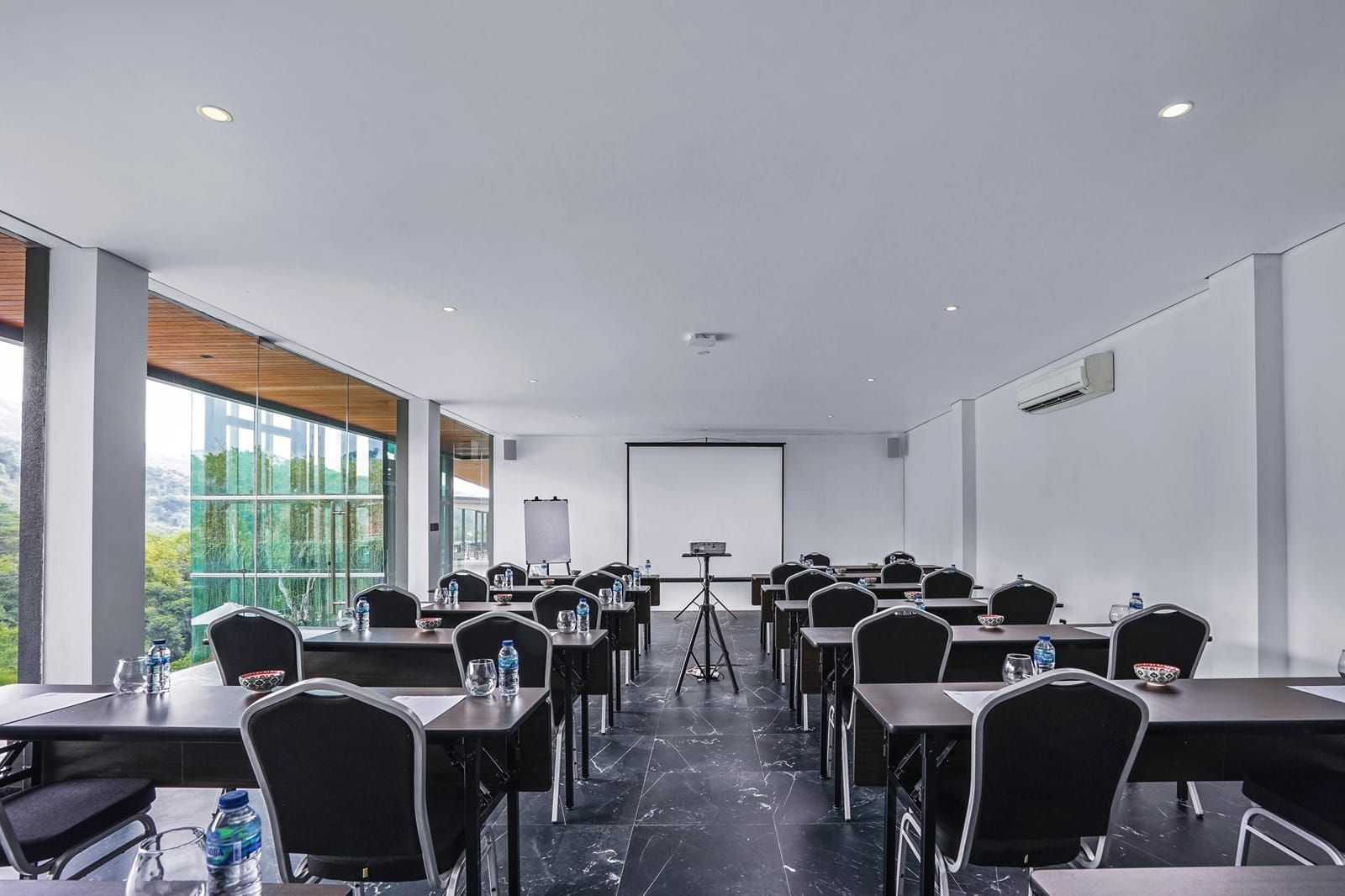 A conference room with tables, chairs, a projector screen, and a large window offering a view of greenery.