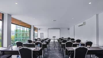 A conference room with tables, chairs, a projector screen, and a large window offering a view of greenery.