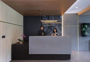 Mostrador de recepción con dos empleados, una pared de mármol y una pantalla de televisión.