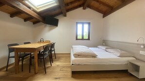 Una habitación con techo de madera, una cama con ropa de cama blanca, una pequeña mesa con sillas y una ventana con vista a la vegetación.