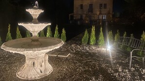 Una fuente de piedra con diseño escalonado, rodeada de grava y pequeños árboles. En el fondo, se observa un edificio con varias ventanas y un balcón.
