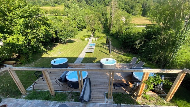 Una terraza de madera con jacuzzis y sillones de descanso, con vistas a un área verde con árboles y cielo despejado.