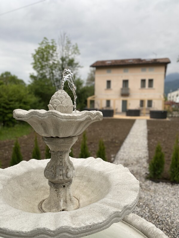 A stone fountain with water spouting from the top, set in a garden with a gravel path leading to a building with a balcony. - Vera Park & Wellness (Belluno)