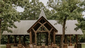 A wooden pavilion with a triangular roof, surrounded by outdoor seating and trees.
