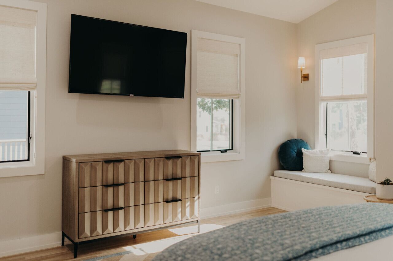 A bedroom with a TV mounted on the wall, a wooden dresser, a window seat with cushions, and a bedside table with a lamp.