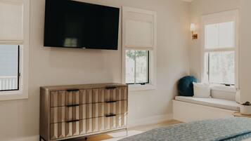 A bedroom with a TV mounted on the wall, a wooden dresser, a window seat with cushions, and a bedside table with a lamp.
