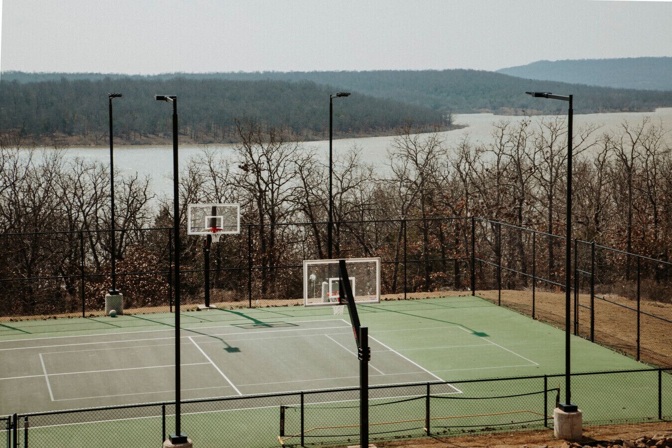 An outdoor basketball court with a net, surrounded by a fence, and overlooking a body of water with trees in the background.