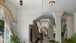 Un vaste hall aux murs blancs, orné de colonnes et doté d’un sol en marbre. On y trouve des plantes en pot et un escalier menant à un autre espace.