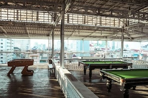 Zona de piscina interior con varias mesas de billar y un banco de madera, con vistas a la ciudad a través de amplias ventanas.