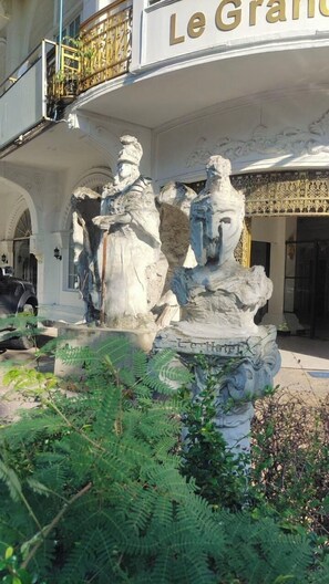 Una fontana con sculture classiche di fronte a un edificio con un cartello che riporta la scritta 'Le Grand'.