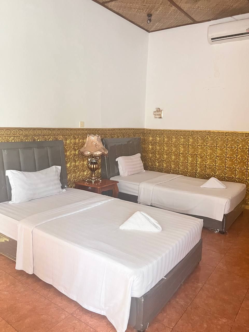 Habitación de hotel con dos camas individuales, una mesita de noche con lámpara y un panel decorativo en la pared.