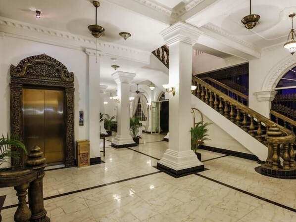 Een statige lobby met een marmeren vloer, een centrale trap en een grote, versierde lift.