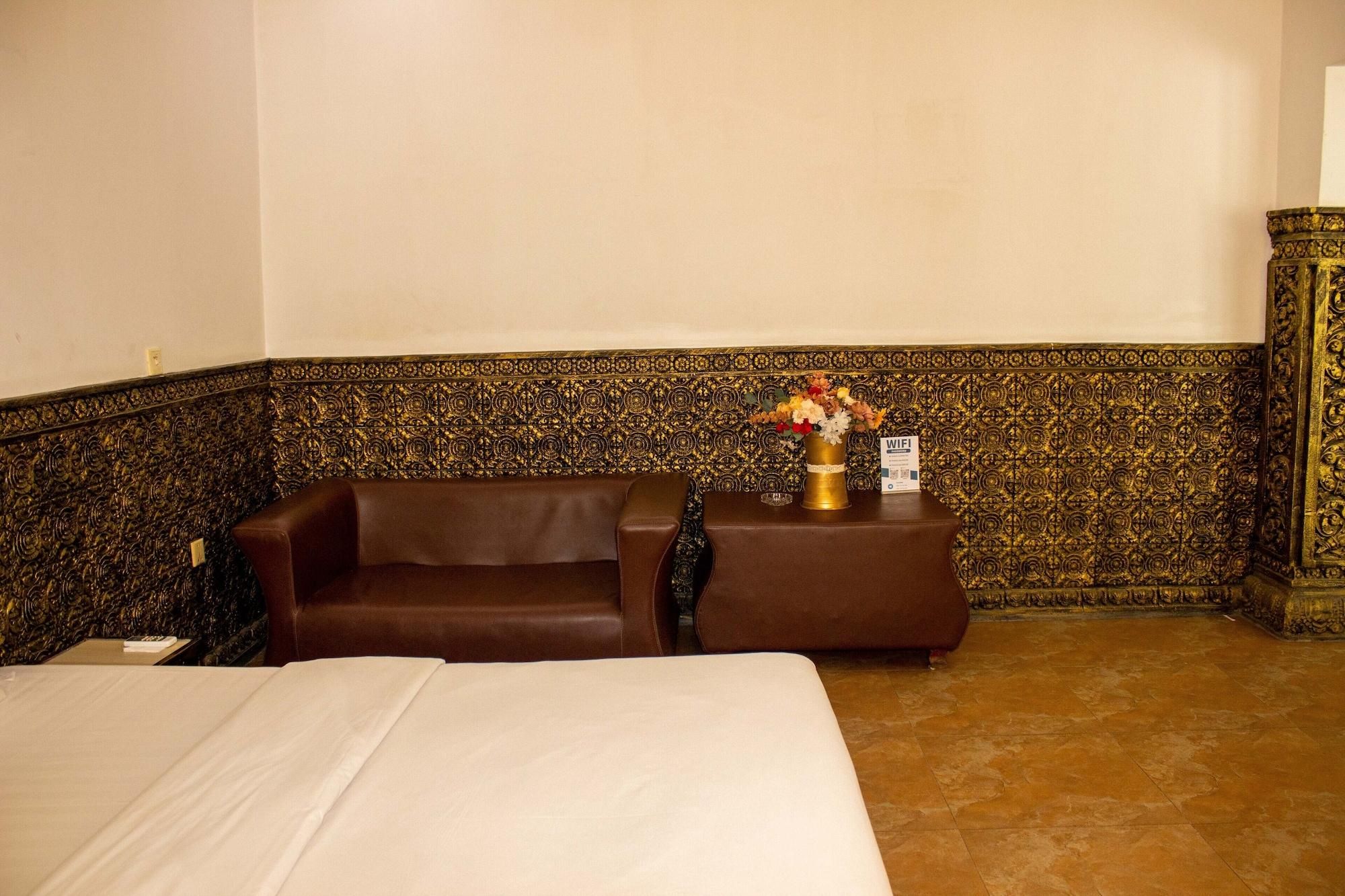 Een hotelkamer met een bed, een bruine bank, een klein tafeltje met een bloevas en een decoratieve muur met ingewikkelde patronen.