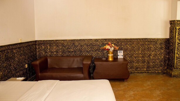 Een hotelkamer met een bed, een bruine bank, een klein tafeltje met een bloevas en een decoratieve muur met ingewikkelde patronen.