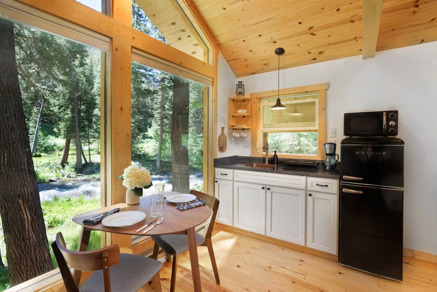 Une cuisine de style rustique avec une table à manger, des chaises, un réfrigérateur noir et une vue sur une forêt grâce à de grandes fenêtres.