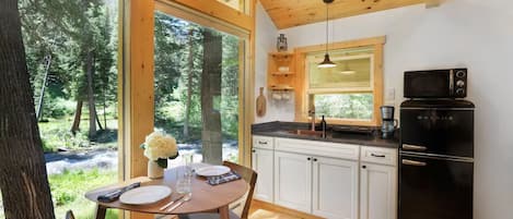 Cocina de cabaña con mesa de comedor, sillas, un refrigerador negro y vista a un bosque a través de amplias ventanas.