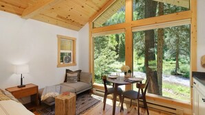 A cozy room with a wooden ceiling, a sofa, a dining table with chairs, and large windows offering a view of a forest. - Creekside Maroon Bells Cabin at T-Lazy-7 Ranch, 5 Min to Maroon Bells & Highlands, 10 to Aspen (Aspen)
