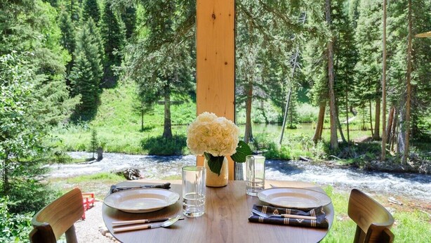 A dining table set with plates, cutlery, and glasses, with a vase of flowers in the center, overlooking a forested area. - Creekside Maroon Bells Cabin at T-Lazy-7 Ranch, 5 Min to Maroon Bells & Highlands, 10 to Aspen (Aspen)