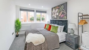 A modern bedroom with a large bed, a headboard with a floral painting, a bedside table with a lamp, and a window with a view of buildings.