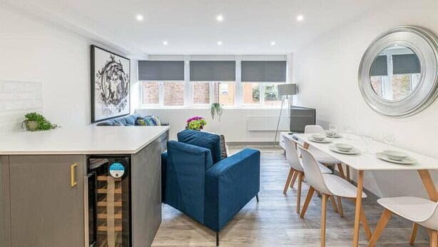 A modern kitchen with a built-in dishwasher, a dining area with a white table and chairs, and a large window with blinds.