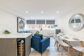 A modern kitchen with a built-in dishwasher, a dining area with a white table and chairs, and a large window with blinds. - Charles House by Guestz (Preston)
