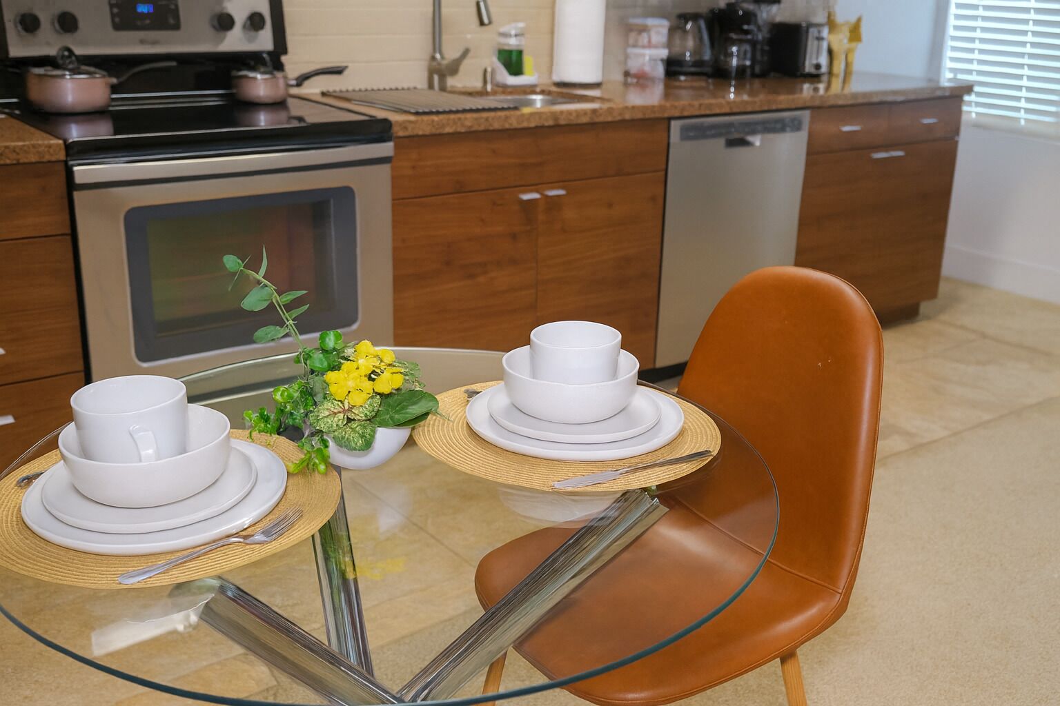 A modern kitchen with a glass table set for two, a brown leather chair, and a stove with pots.
