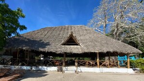 Una cabaña con techo de paja y una ventana triangular, rodeada de arena y asientos de madera.