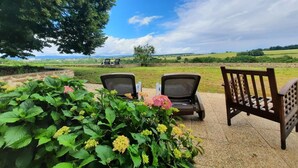 Un espace extérieur aménagé avec deux chaises et une table, entouré d’une végétation luxuriante et offrant une vue sur un paysage rural.