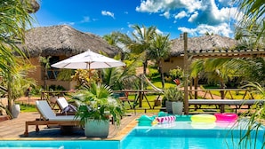 Un área de piscina con sillas de descanso, una estructura de techo de paja y plantas en macetas.