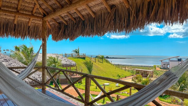 Una hamaca bajo techo de paja, con vista a una playa y un jardín cuidado.