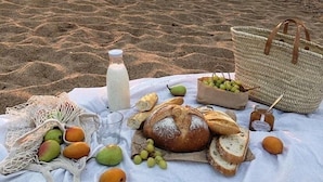 Un montaje para picnic en la playa, con una manta, pan, frutas, una botella y una cesta.