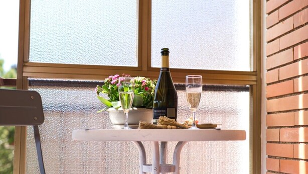 Et bord med en flaske Prosecco, et vinglas og en skål med snacks på en balkon med udsigt til en murstensvæg og et vindue.