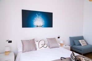 Un dormitorio con una cama, un sillón azul, una mesita de noche con una lámpara y una obra de arte en la pared que representa un chapuzón de agua.