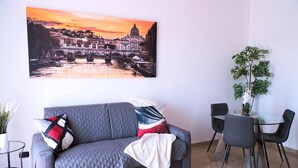 En stue med en grå sofa, et glasbord med stole, en potteplante og et stort bybillede på væggen.