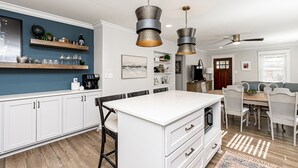 Una cocina moderna con armarios blancos, una isla central, lámparas colgantes y un comedor con sillas blancas.