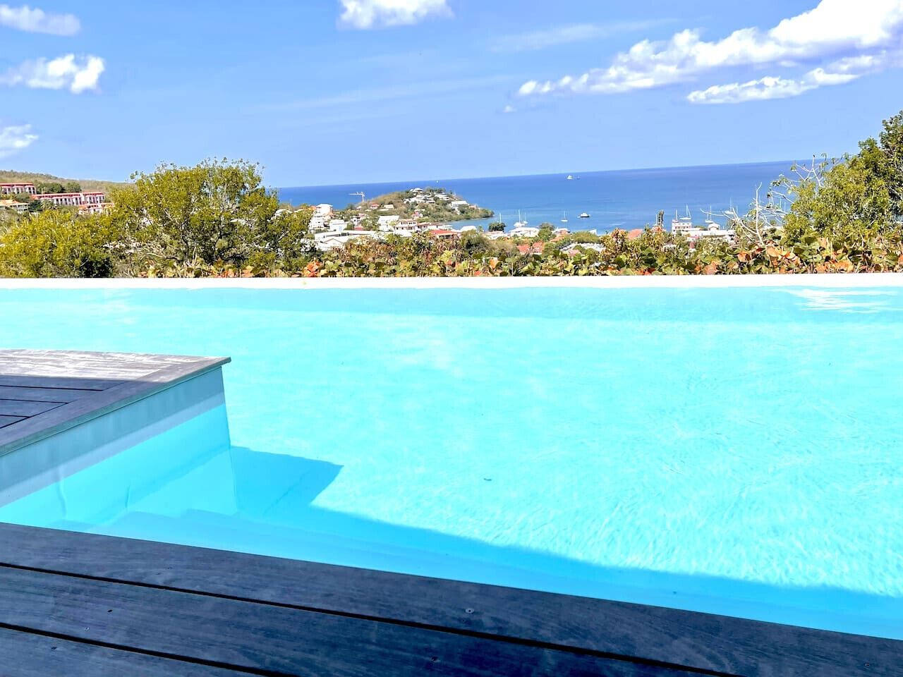 Une piscine d’un bleu éclatant, bordée d’une terrasse en bois, qui offre une vue sur une ville côtière et la mer.