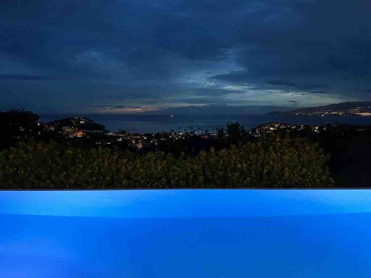 Une vue nocturne d’une ville côtière, avec des bâtiments et une étendue d’eau en arrière-plan.