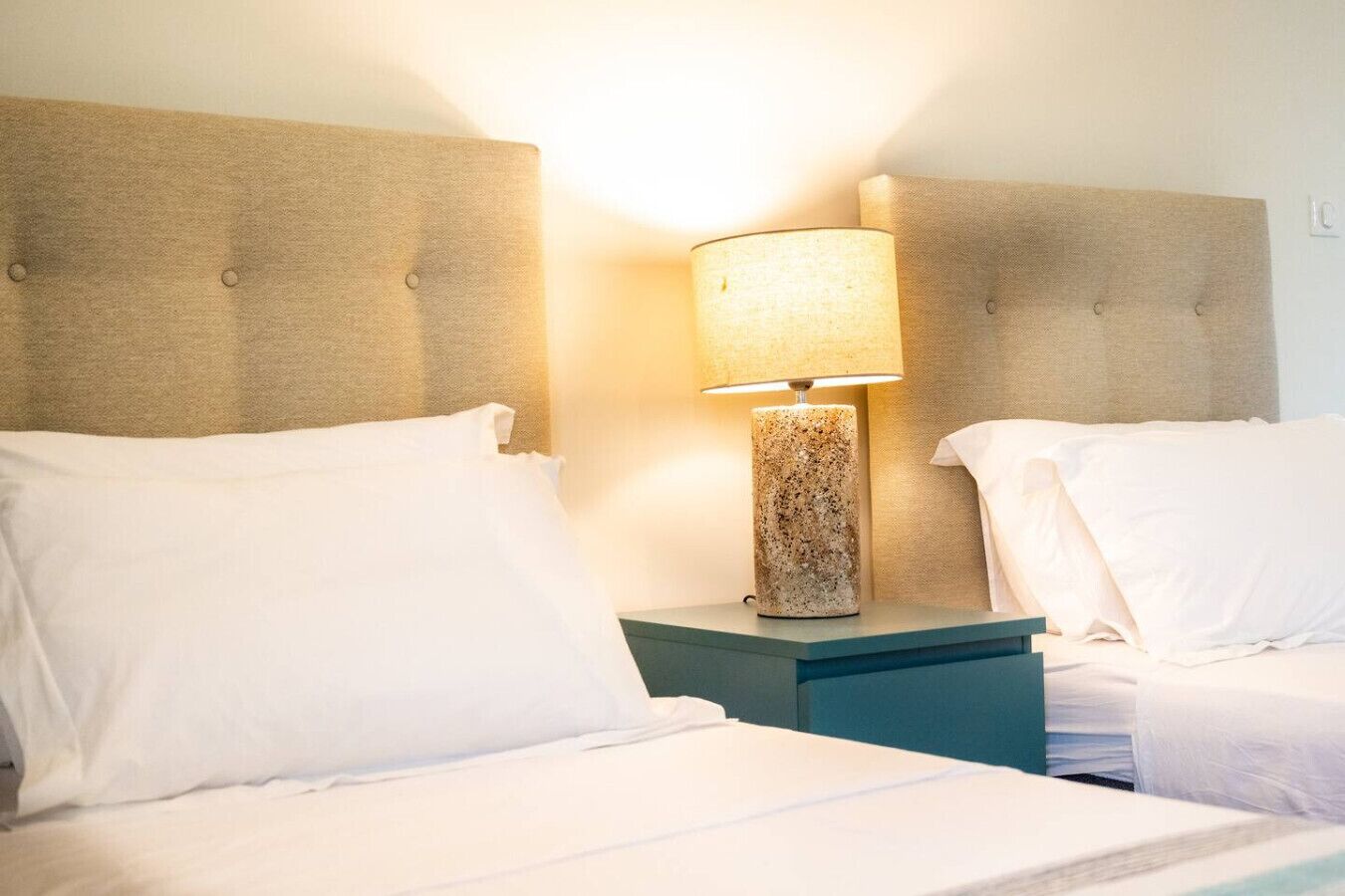 A hotel room with two beds, each with white linens and pillows, and a nightstand with a textured lamp between them.