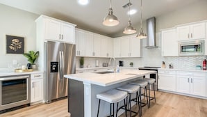 A modern kitchen with white cabinets, stainless steel appliances, a large island with bar stools, pendant lighting, and a microwave.