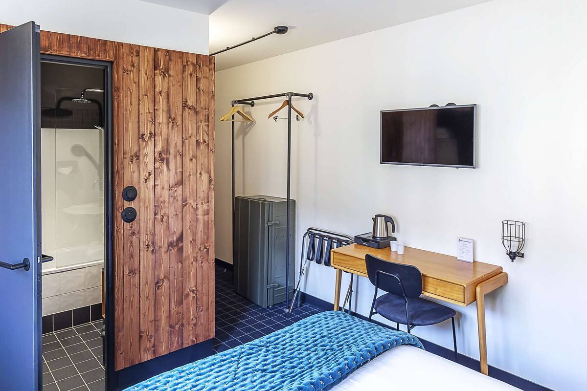 A compact room with a wooden wardrobe, a bed, a desk with a chair, a television, and a shower area.