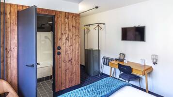 A compact room with a wooden wardrobe, a bed, a desk with a chair, a television, and a shower area.