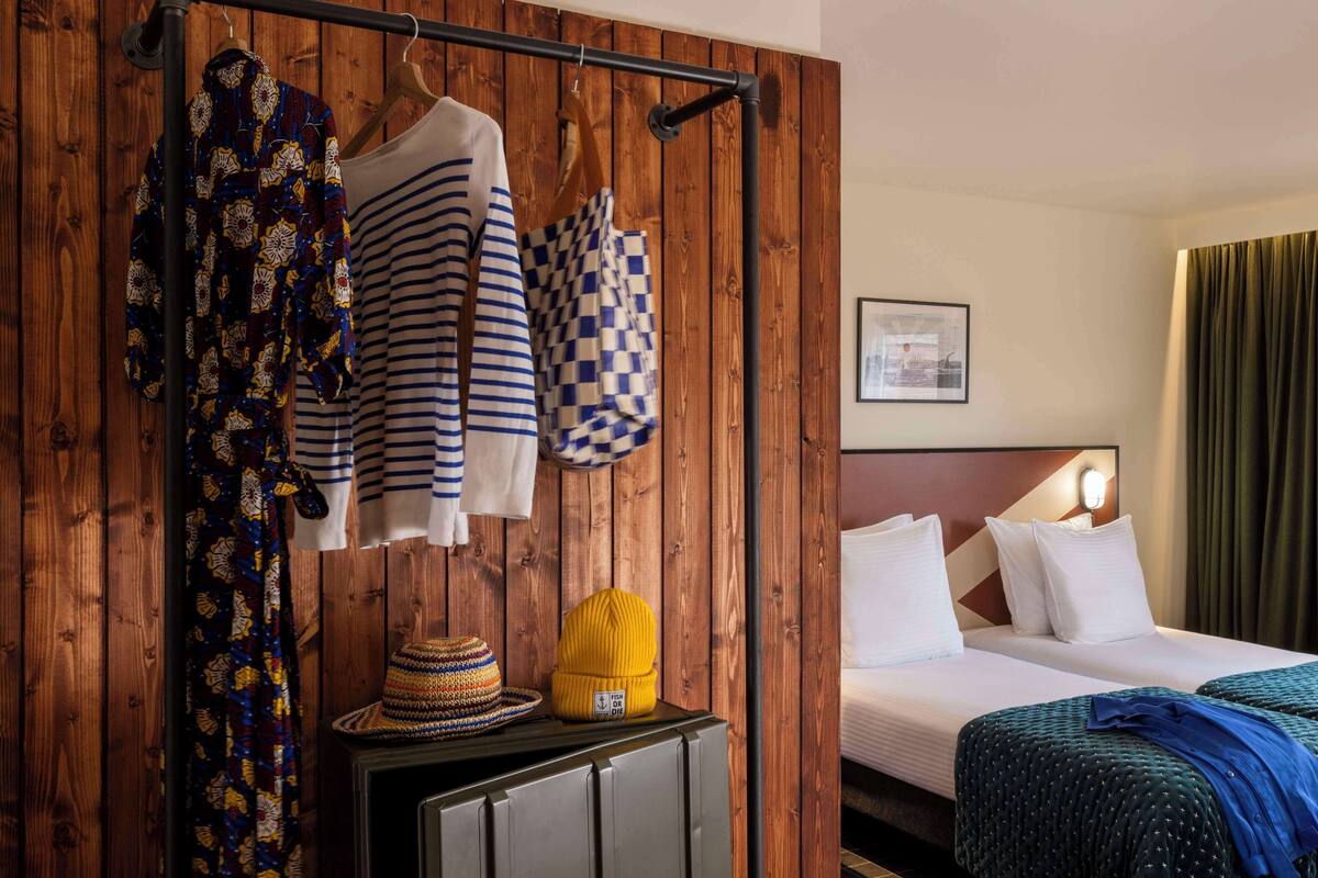 A hotel room with a wooden wall, a bed with white linens, a striped shirt, a checkered bag, a yellow beanie, and a woven hat.